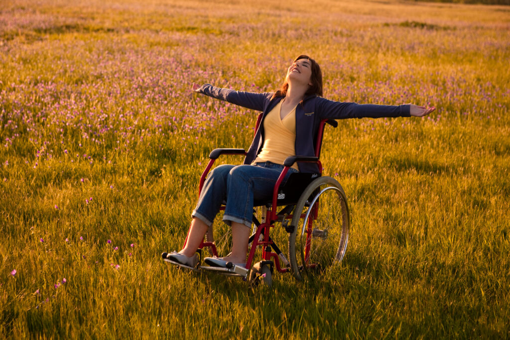 Glad kvinna i rullstol med armarna utsträckta. På gräsyta med blommor.