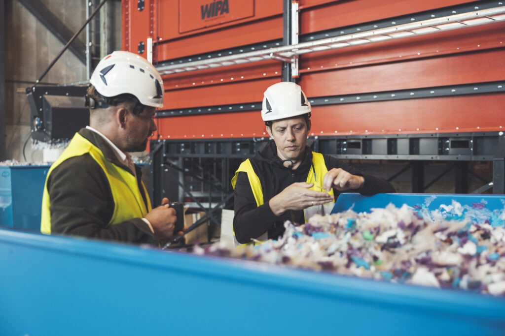 Två personer i skyddskläder och hjälmar står i en container med plast.