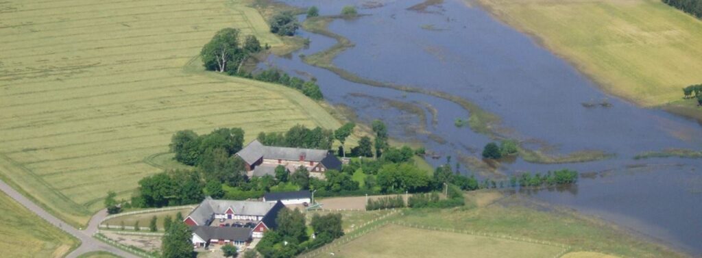 Flygbild över kringbyggda gårdar, åkrar samt ett översvämmat vattendrag.