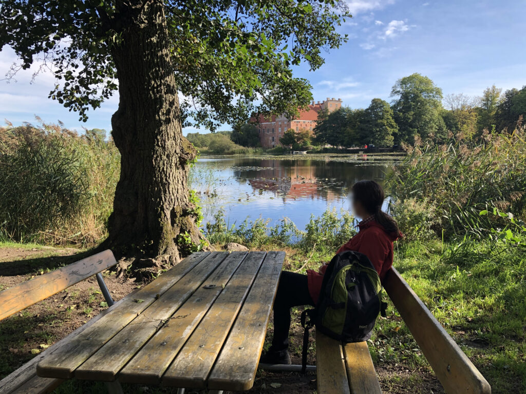 En människa sitter på en träbänk vid ett träbord i naturen. I bakgrunden en sjö och ett slott.
